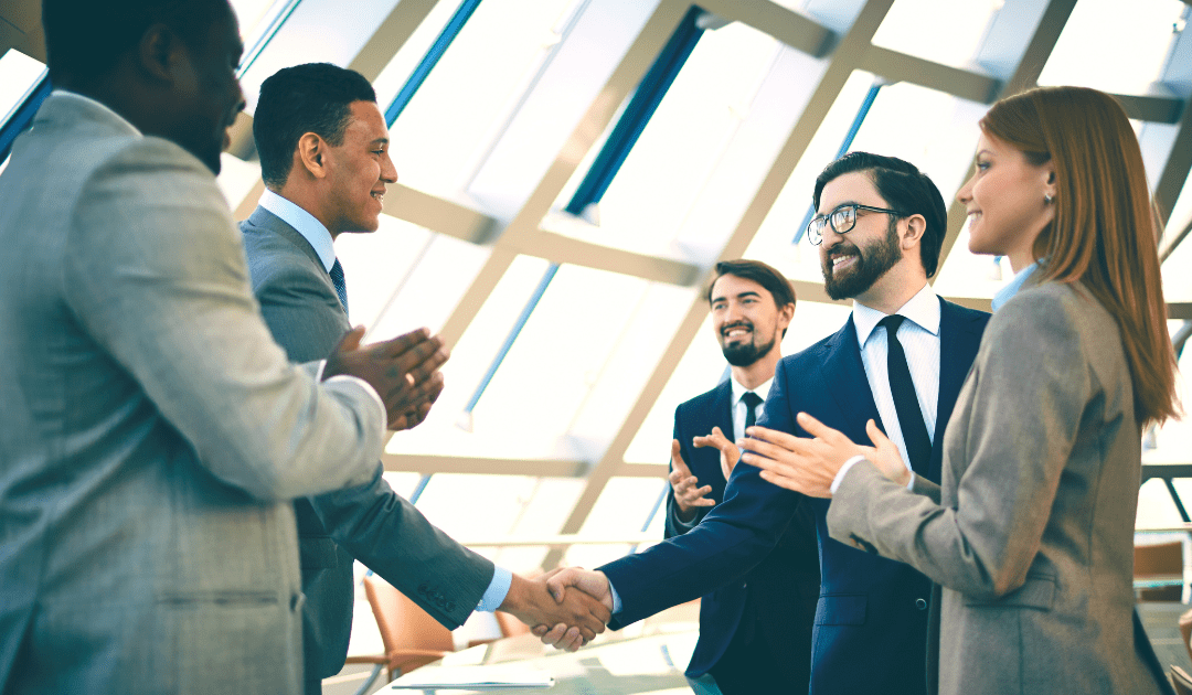 Four professionals in business attire are engaged in a discussion in a modern office setting with large windows. Two men are shaking hands while another man and a woman stand nearby clapping and smiling, indicating a celebratory or congratulatory moment.