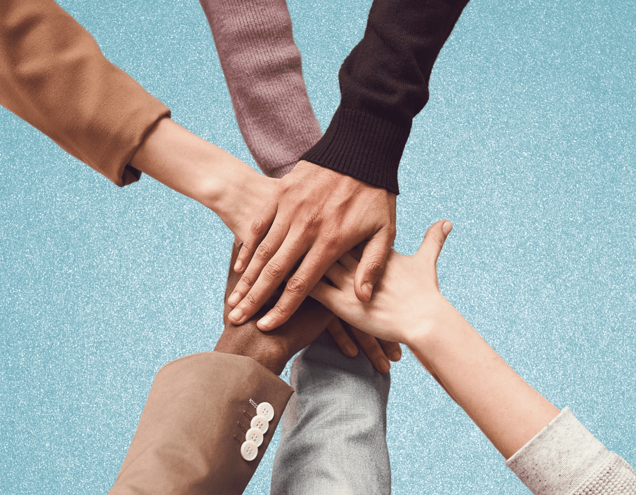 A diverse group of six hands, belonging to people of different skin tones and genders, join together in a supportive gesture against a light blue background. Each person's arm is dressed differently, highlighting a mix of styles and colors.