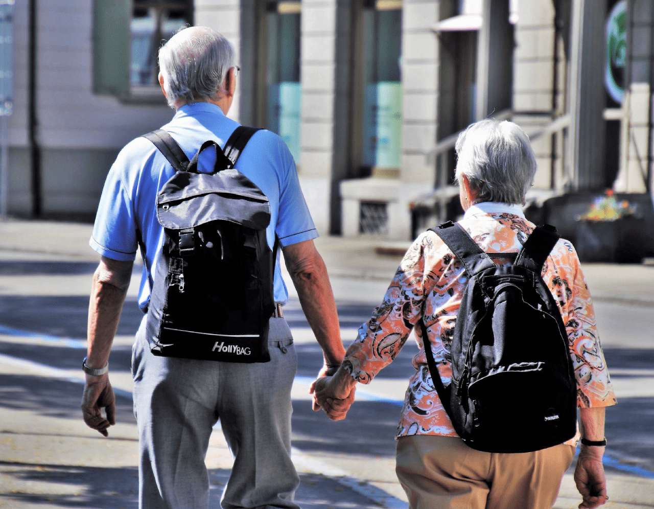 An elderly couple walking hand in hand on a sunny sidewalk. Both have gray hair and are wearing backpacks. The man is wearing a blue polo shirt and gray pants, and the woman is wearing a floral blouse and beige pants. They are seen from behind.